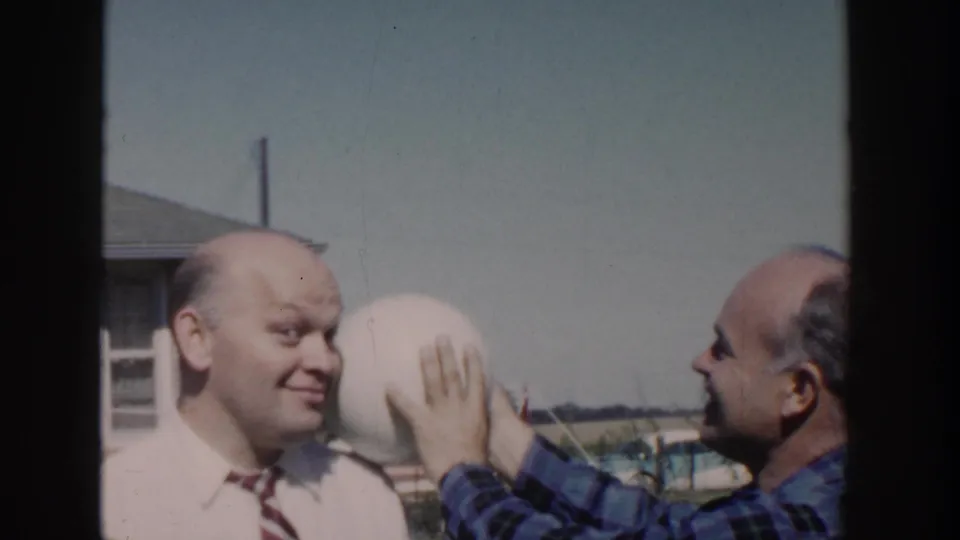 1958 Wisconsin backyard frame showing a man in plaid holding a pale round object against a smiling bald man's head during a comic bit.