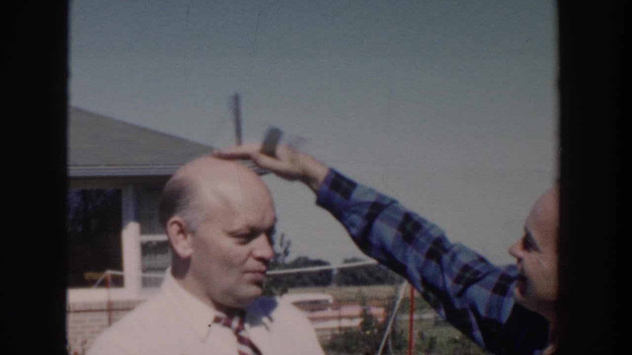 In a 1958 Wisconsin backyard scene, a man in a blue plaid shirt pats the head of a bald man in a white shirt and striped tie.