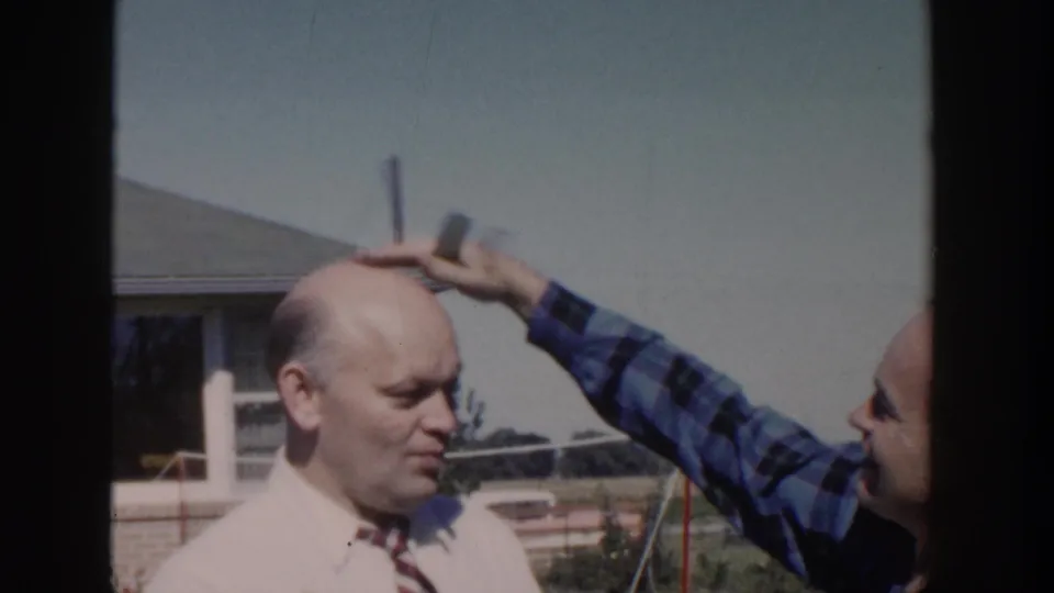 In a 1958 Wisconsin backyard scene, a man in a blue plaid shirt pats the head of a bald man in a white shirt and striped tie.