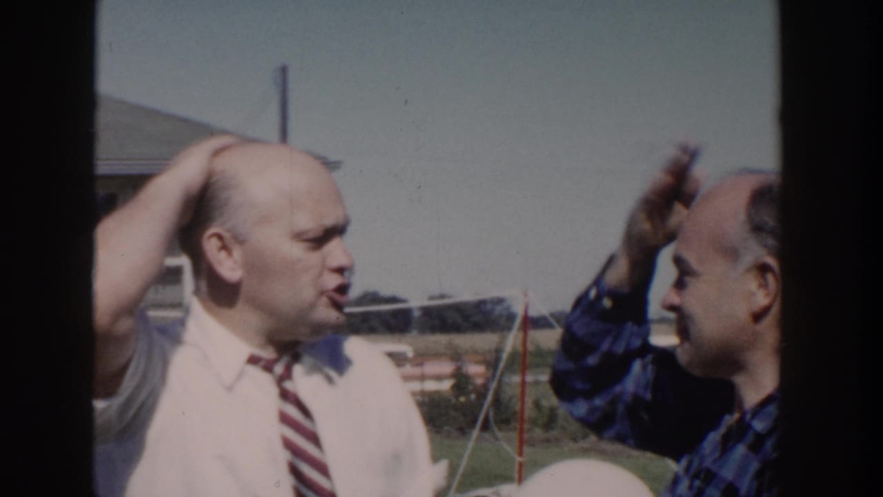 Bald man in shirt and tie speaks while another man raises a hand near his head.