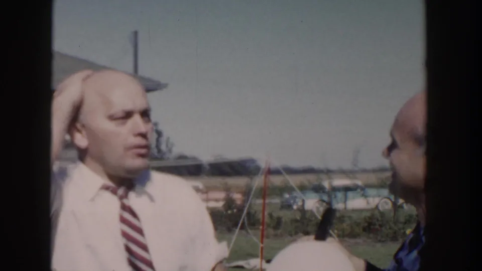 In the sunny Wisconsin yard, the man in the white shirt and tie reacts as another bald man stands close beside him.