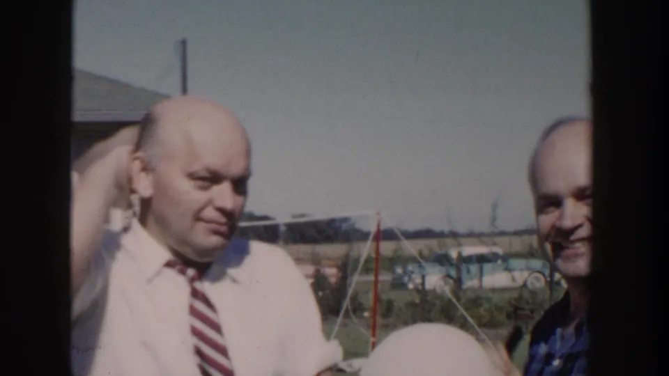 The man in the white shirt smiles while the man in the blue plaid shirt grins beside him in the 1958 Wisconsin backyard footage.
