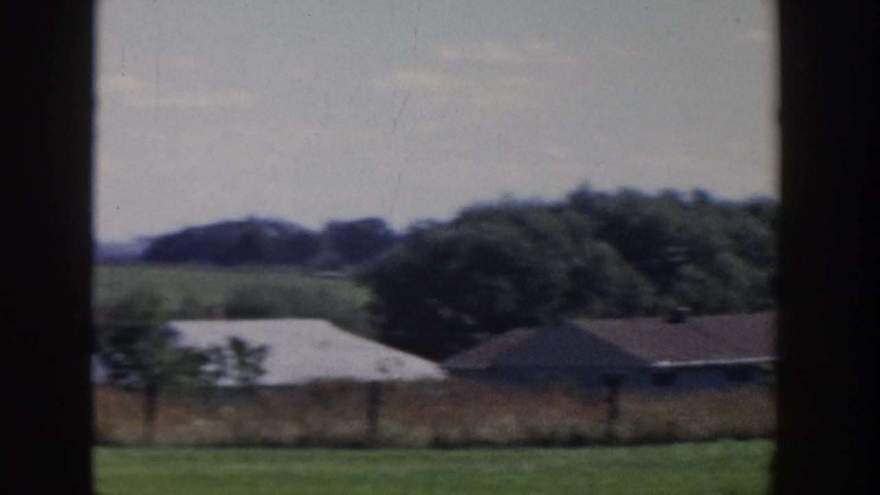 1958 Wisconsin rural landscape with fields, low farm buildings, tree line, and utility poles viewed through a dark window frame.