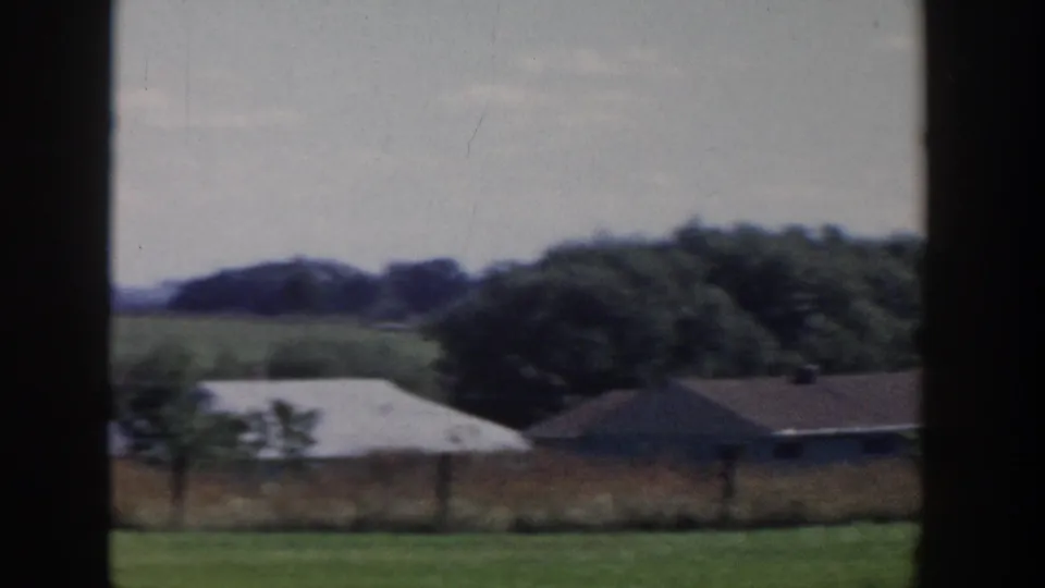 1958 Wisconsin rural landscape with fields, low farm buildings, tree line, and utility poles viewed through a dark window frame.