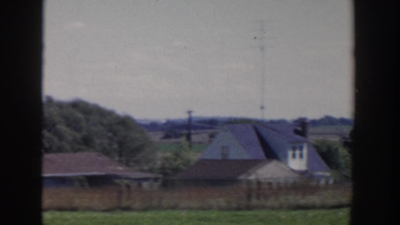 Farmhouse roofs, trees, fields, and utility poles beyond a dark frame.