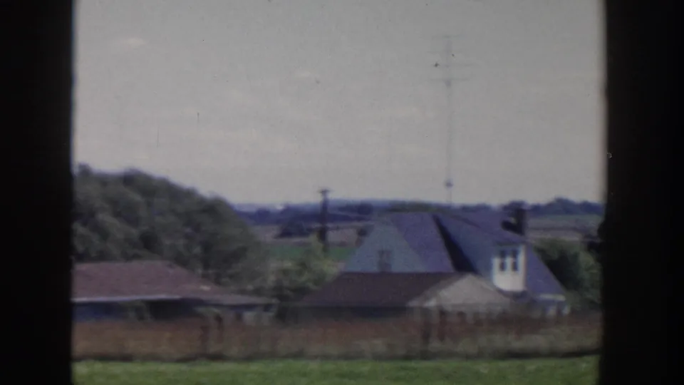 Rural Wisconsin view with sloped-roof houses or farm buildings, fields, trees, and utility poles under a pale sky.