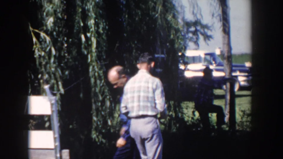 1958 Wisconsin home-movie frame showing two adult men near a wire fence and wooden structure, with a light-colored car behind them.