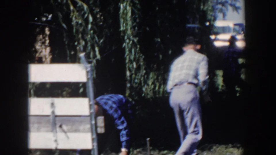 1958 Wisconsin frame with one adult man bending near a wooden structure and another walking away beside willow branches and a fence.