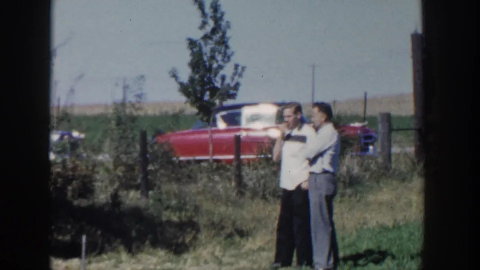 Two adult men stand close together in a grassy rural area, one appearing to smoke, with a red convertible and fence line behind them.