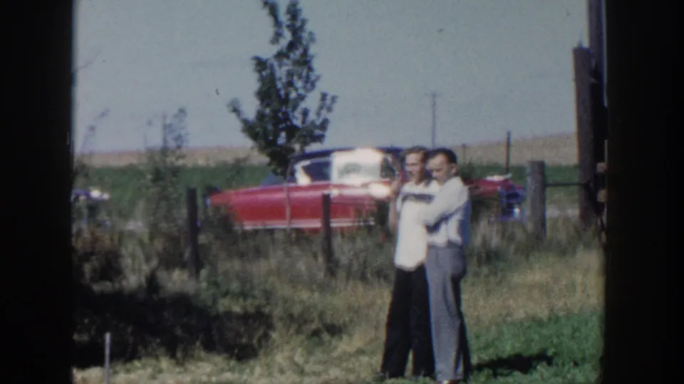A 1958 Wisconsin home-movie frame shows two adult men standing in grass, with a red 1950s convertible, fence posts, and open land behind them.
