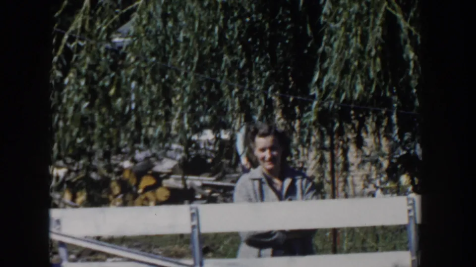 In Wisconsin 1958 home-movie footage, a woman stands behind a white fence in a shaded yard with willow branches overhead.