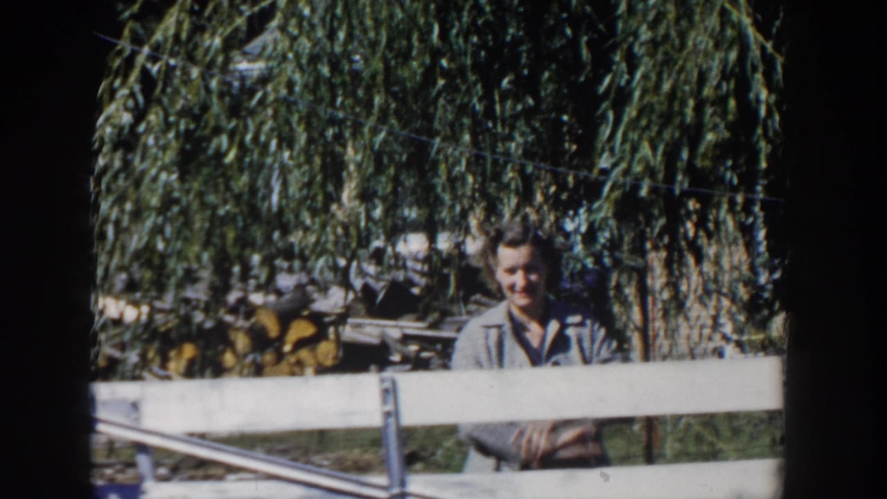 A woman smiles toward the camera from behind a white fence in a Wisconsin yard, with willow branches and stacked wood in the background.