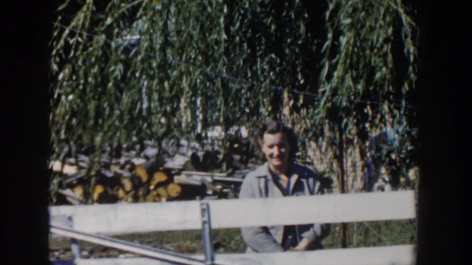 The woman remains behind the white fence under willow shade, with soft 8mm focus and a yard backdrop of grass and cut wood.