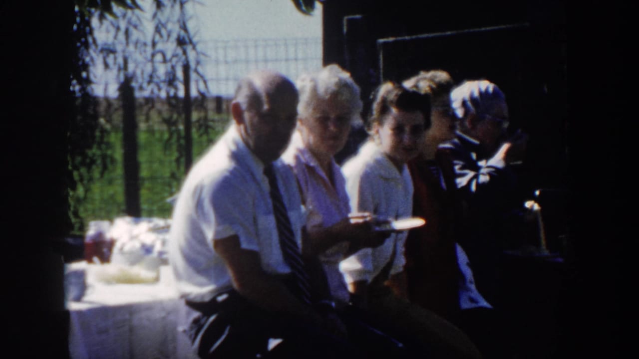 Five adults sit close together outdoors in Wisconsin home-movie footage, holding plates beside a dark doorway and sunlit grass.
