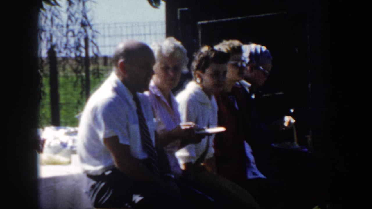 The group remains seated in strong shade, with a man in a white shirt and tie at left and sunlit grass behind.