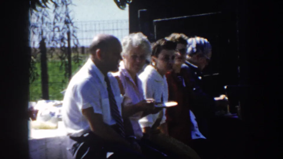 Adults sit shoulder to shoulder on a shaded bench or ledge, with one woman holding a plate and others turned toward the camera.