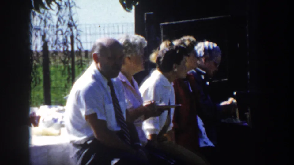 The adults turn slightly along the shaded bench while holding plates, with bright grass and a wire fence visible behind them.