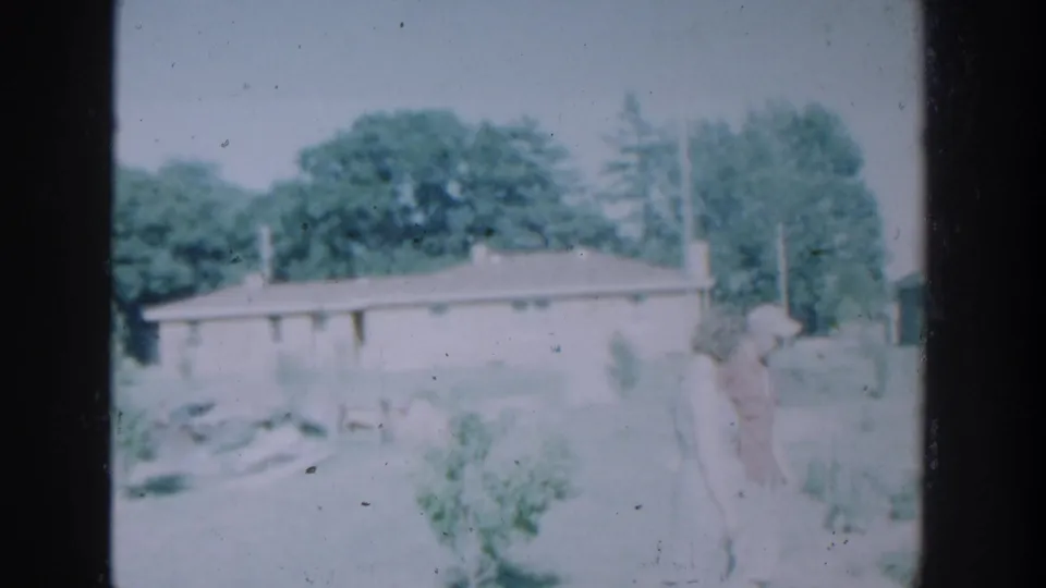 1964 Wisconsin home-movie frame showing two adults in a grassy yard before a single-story house with trees behind it.