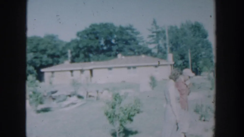 1964 Wisconsin 8mm frame with two adults in a residential yard, a low house, garden plants, and trees in the background.