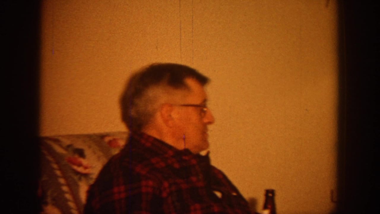 1964 Wisconsin home-movie frame showing a man in glasses seated beside a patterned chair in a dim living room.