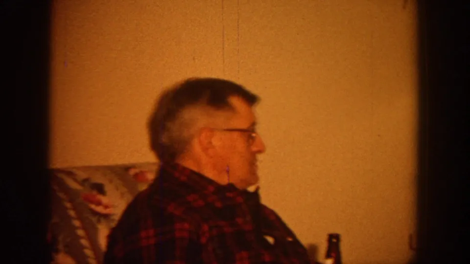 1964 Wisconsin home-movie frame showing a man in glasses seated beside a patterned chair in a dim living room.