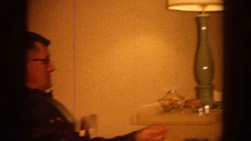1964 Wisconsin home-movie frame with a man in profile, a tall green lamp and a side table in a private living room.