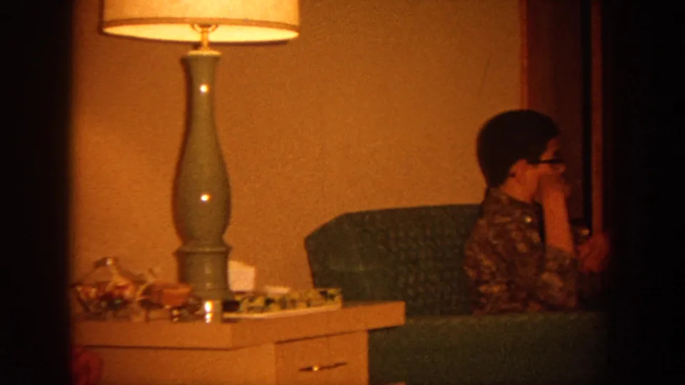 1964 Wisconsin home-movie frame showing a seated woman or teen in a floral dress near a blue-green chair and tall lamp.
