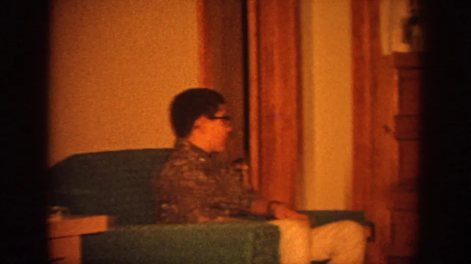 1964 Wisconsin home-movie frame with a seated woman or teen in glasses facing toward the television in a warm private interior.