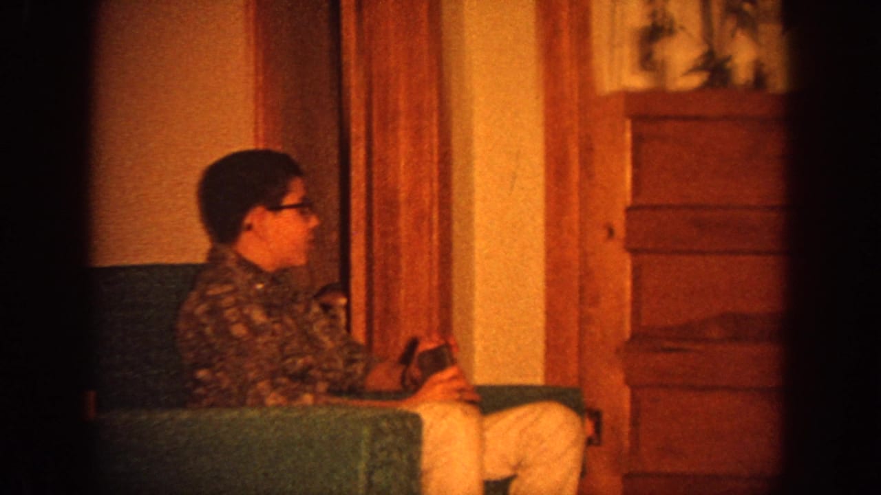 1964 Wisconsin home-movie frame showing a seated person in glasses on a green sofa in a warm living room interior.