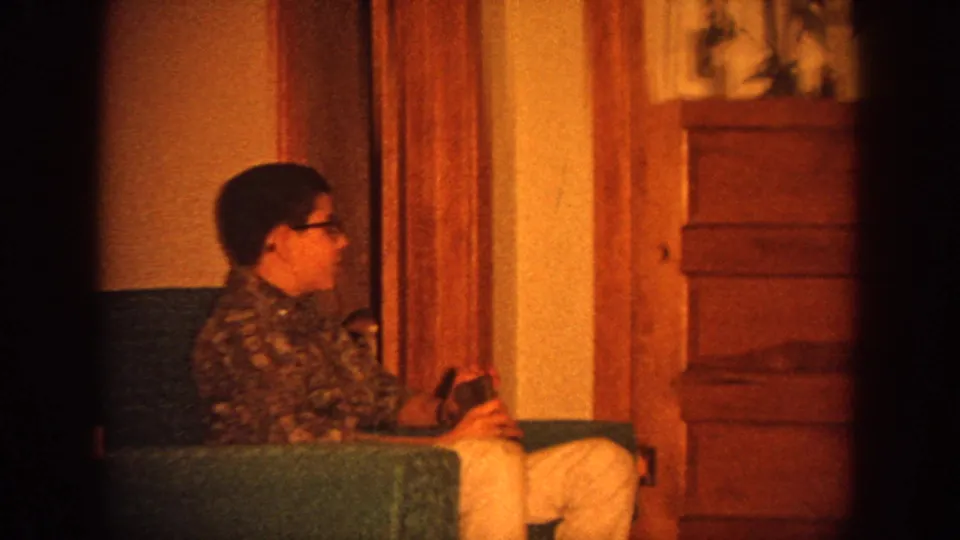 1964 Wisconsin home-movie frame showing a seated person in glasses on a green sofa in a warm living room interior.