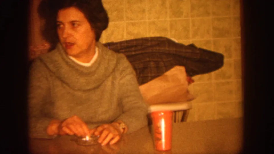 1964 Wisconsin home-movie frame showing a woman in a gray cowl-neck sweater seated at a kitchen table.