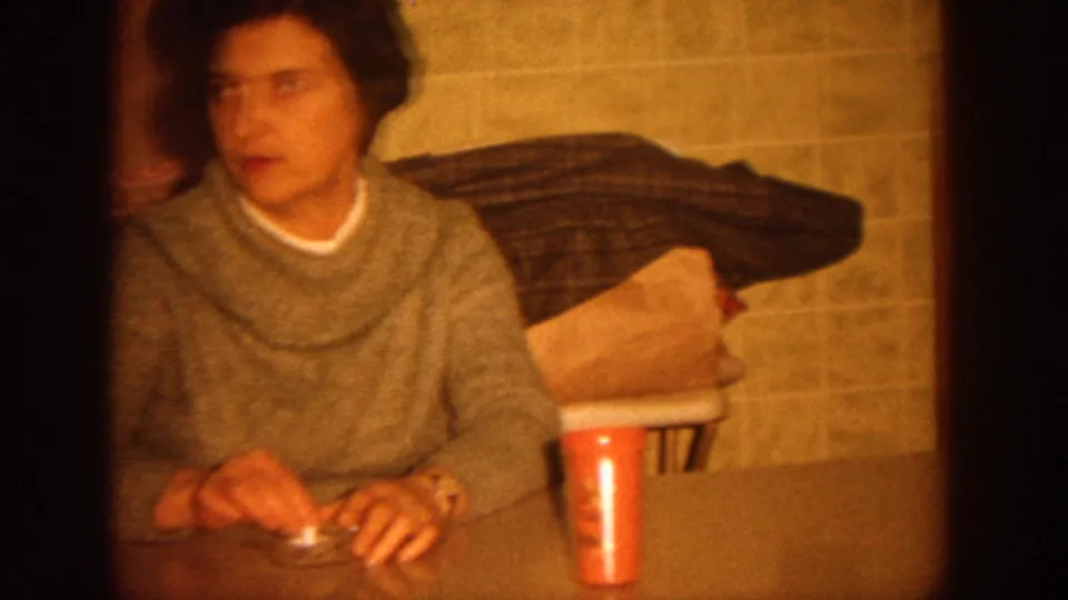 1964 Wisconsin home-movie frame showing a woman at a kitchen table with tiled wall and household items behind her.