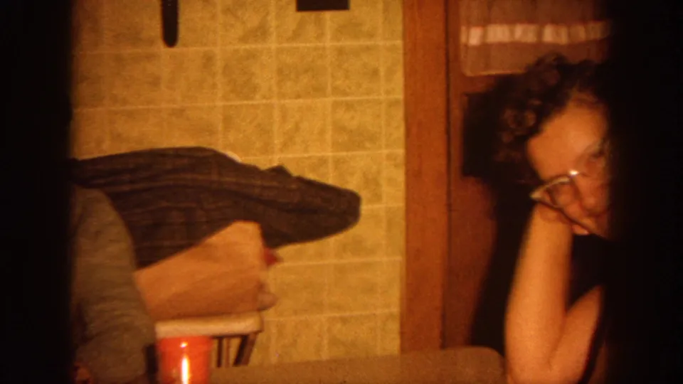 1964 Wisconsin home-movie frame showing a woman with glasses seated beside a kitchen table in a private interior.