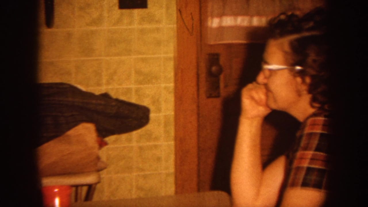 In 1964 Wisconsin home-movie footage, a woman in glasses and a plaid shirt sits beside tiled kitchen walls and wood trim.