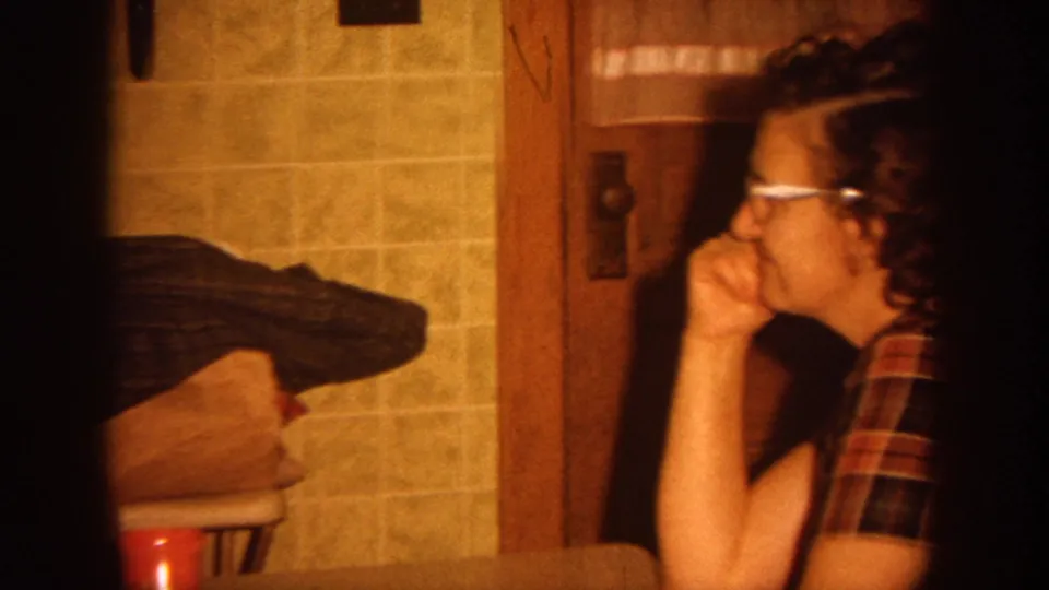 In 1964 Wisconsin home-movie footage, a woman in glasses and a plaid shirt sits beside tiled kitchen walls and wood trim.