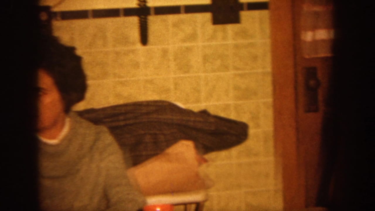 Woman in a gray sweater sits near tiled kitchen walls, partially obscured by a dark film strip edge.
