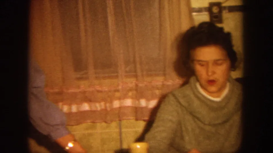 In the lower frame, a woman in a gray sweater looks down while seated near pink curtains and tile in a private Wisconsin interior.