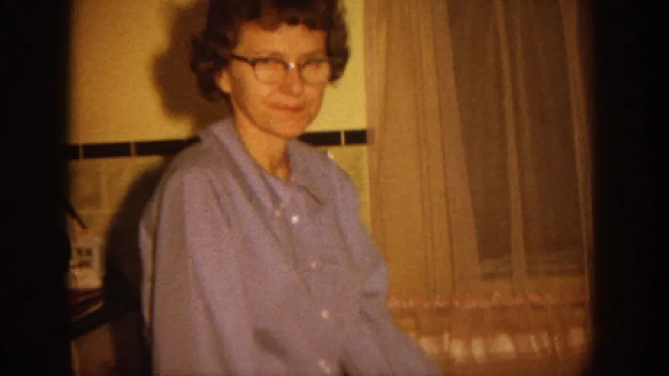 A woman in glasses and a light blue button-up shirt smiles gently in a 1964 Wisconsin kitchen interior with curtains behind her.