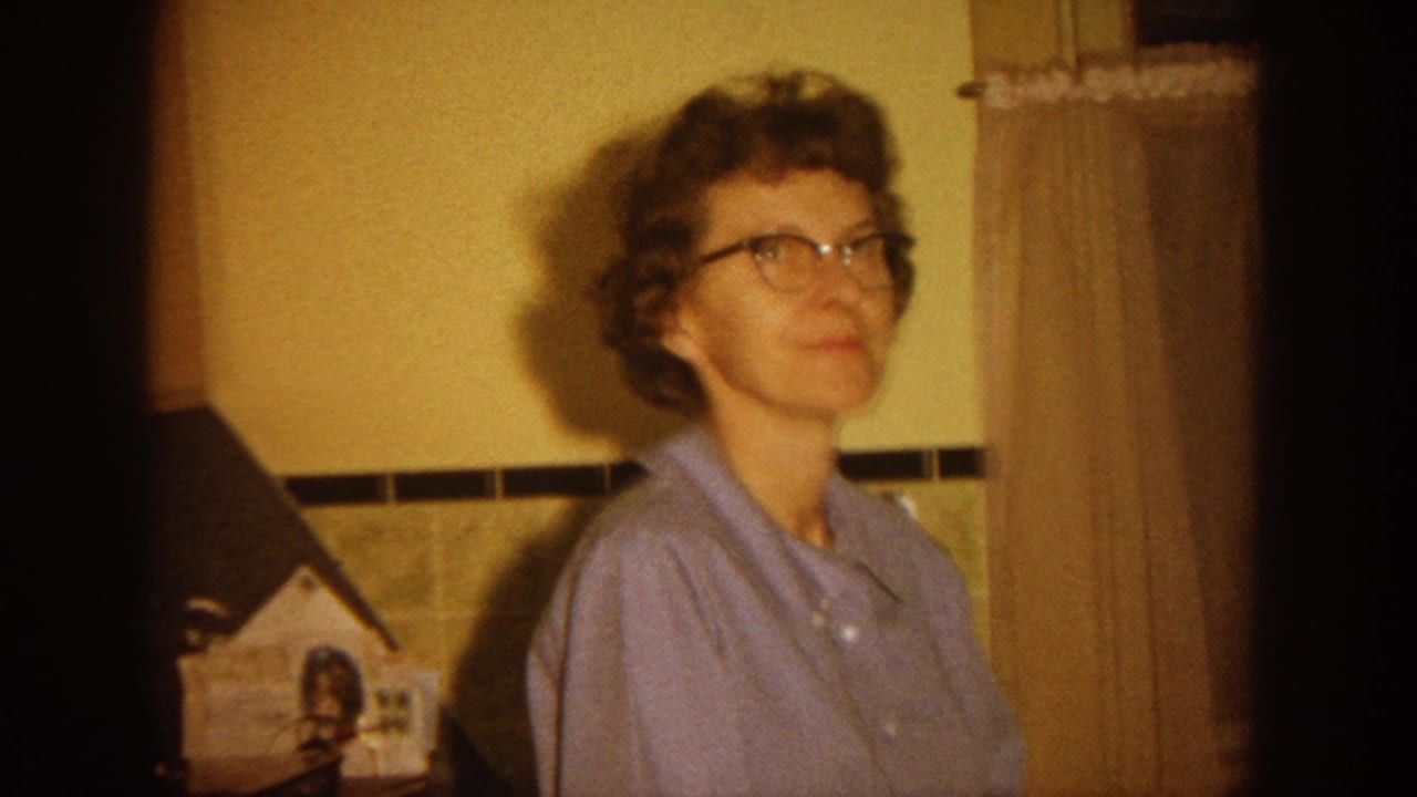 In a 1964 Wisconsin home-movie frame, a woman in glasses and a light blue shirt stands in a warm-lit interior with tiled wall trim.