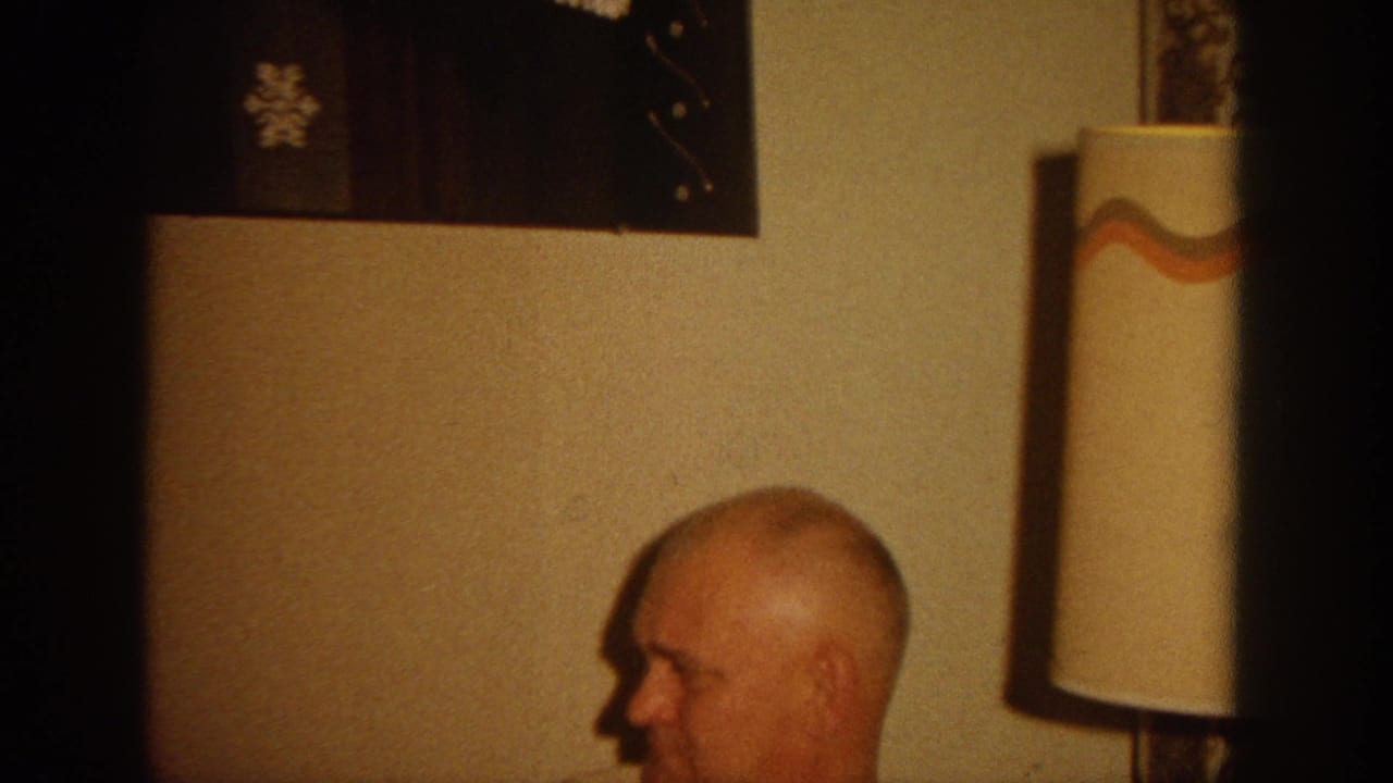 Bald man in profile sits near a wall lamp with a patterned shade.