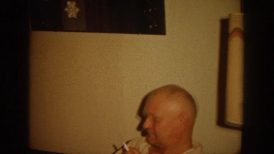 The home-movie frame shows a seated bald man in profile holding a cigarette near wall art and a tall lamp inside a private interior.