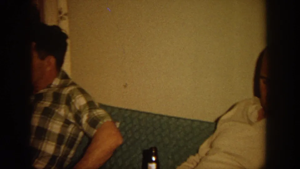 A man in a plaid shirt sits on a green couch in a Wisconsin home interior, with a beer bottle nearby and another person partly visible.
