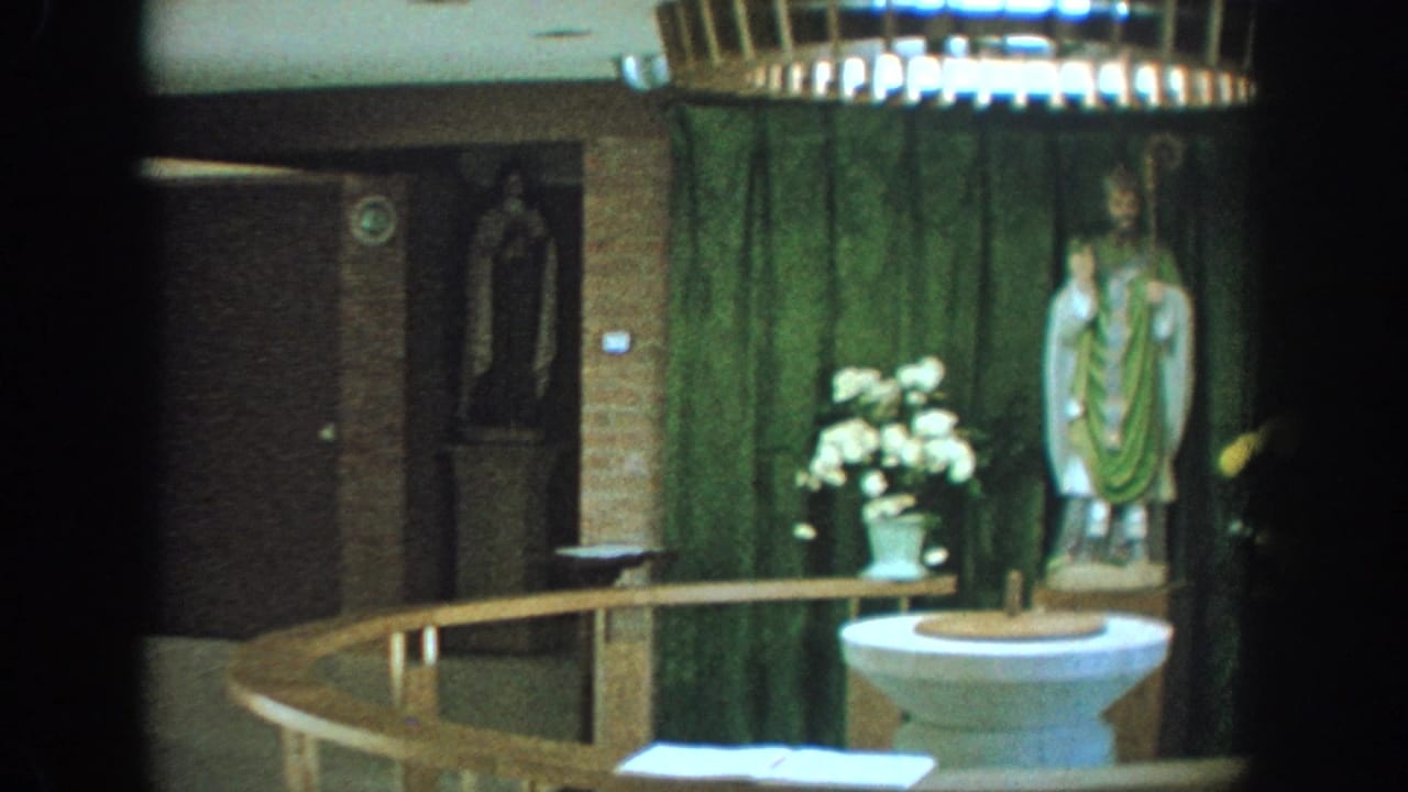1964 Wisconsin 8mm frame showing a church baptismal font area with a saint statue, green curtain, flowers, and wooden rail.