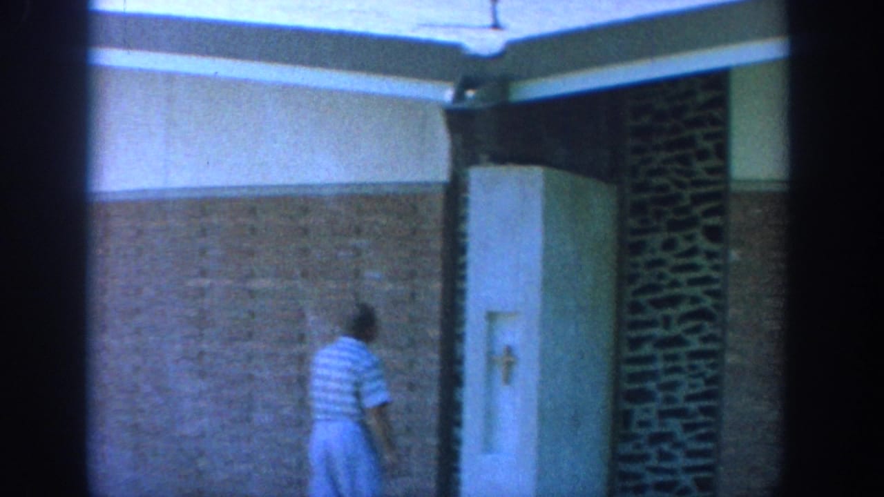 1964 Wisconsin church corridor view with a man walking past textured brickwork and a recessed wall niche bearing a cross.