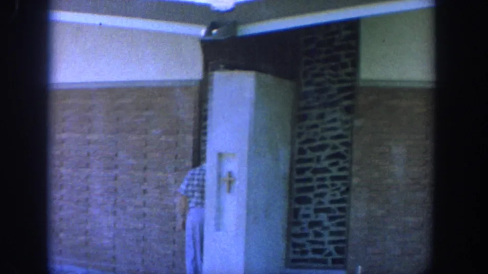 1964 Wisconsin 8mm frame of a man from behind near a brick corridor wall and recessed church niche with a cross.