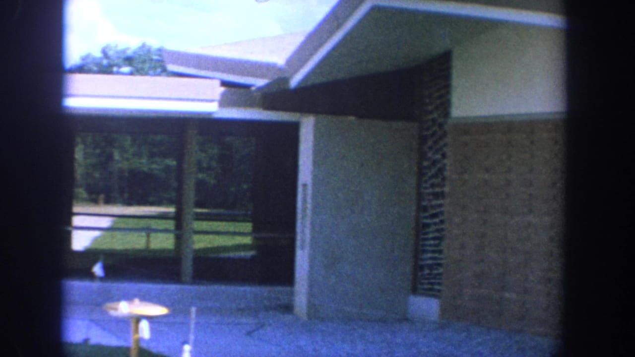 1964 Wisconsin home-movie frame showing a brick-and-glass church exterior with eaves, lawn, sidewalk, and a partially visible woman.