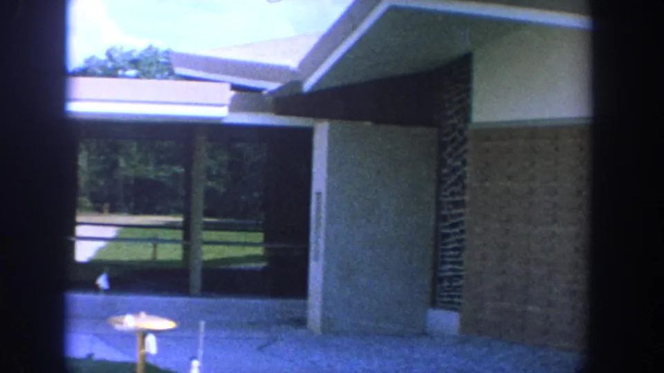 1964 Wisconsin home-movie frame showing a brick-and-glass church exterior with eaves, lawn, sidewalk, and a partially visible woman.
