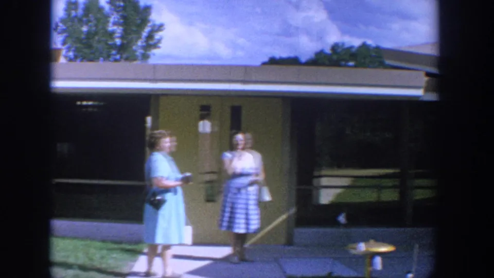1964 Wisconsin frame of two women on a church walkway, one in a light blue dress and one in a blue-and-white checkered skirt.