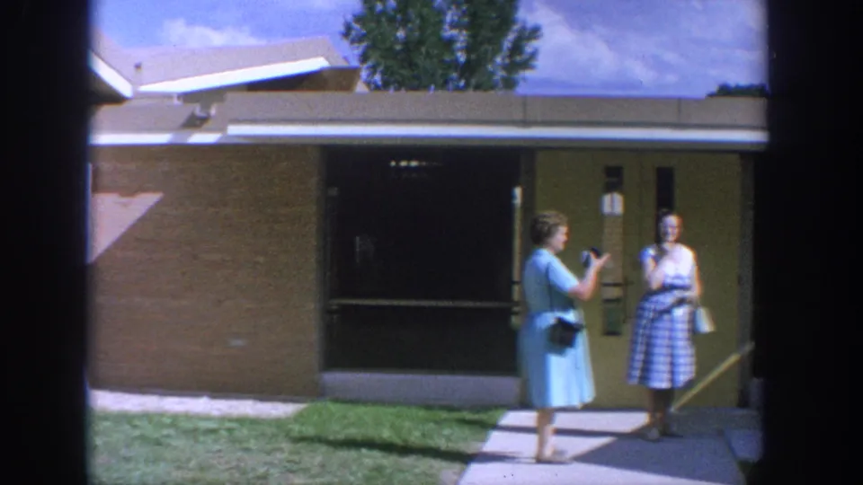 A woman in a blue dress raises a camera toward another woman standing outside the Wisconsin church entrance in 1964.