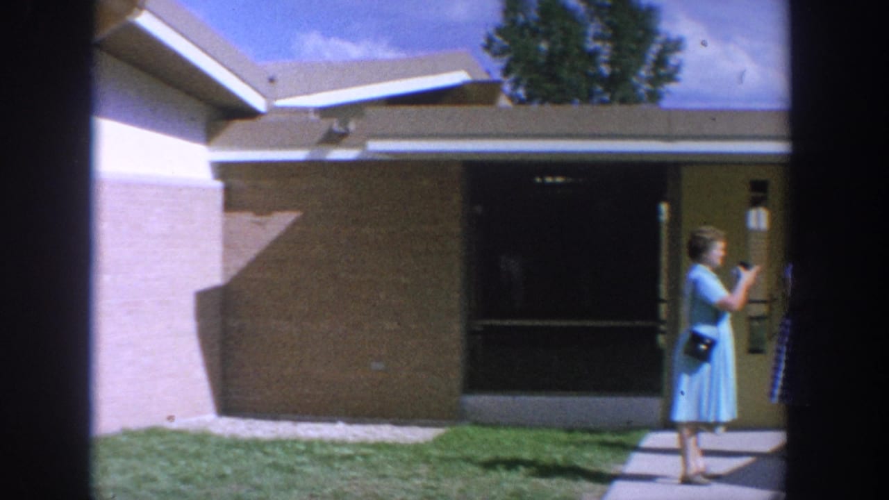1964 Wisconsin 8mm frame showing two women outside a low mid-century building as a man stands near an entrance.